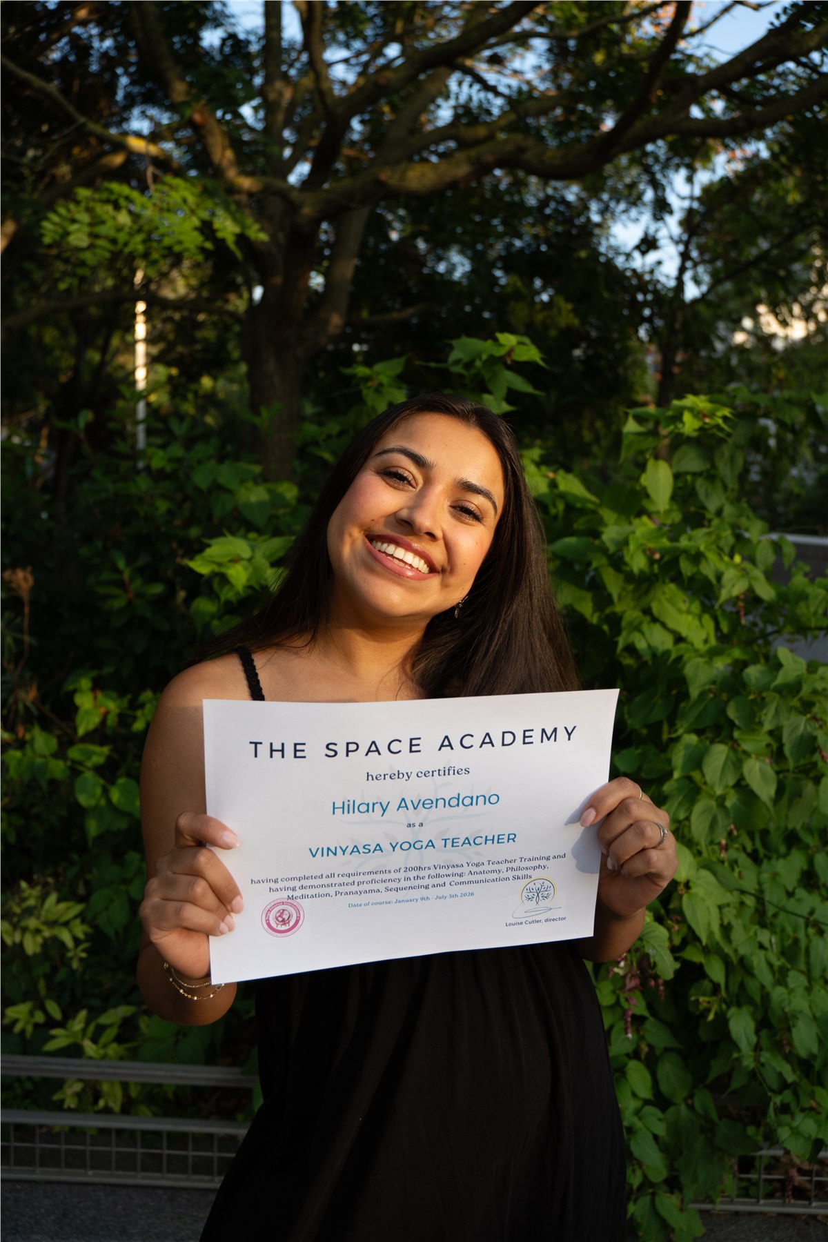 Hilary smiling and holding her yoga teacher training certificate from The Space Academy